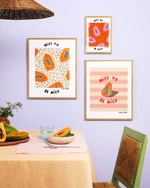 Poster Papaya No1, part of a gallery wall with Poster Papaya and Papaya No3, all in oak frames. Above a dining table set with papayas and an orange tablecloth. A green chair is positioned on one side.