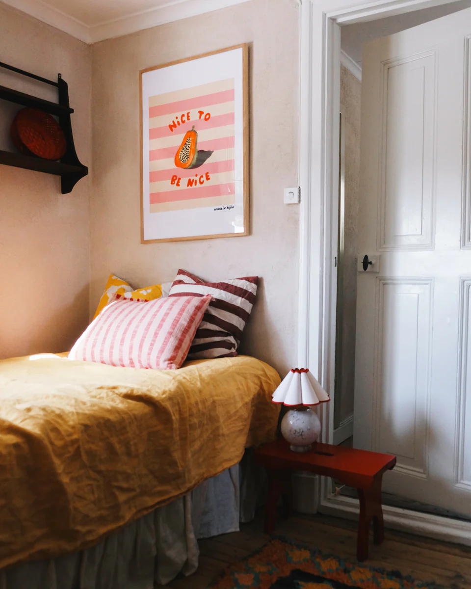 Poster Papaya in oak frame hanging above a bed dressed with linen textiles. A small red stool stands next to the bed with a cute lamp on top. The door is slightly ajar.