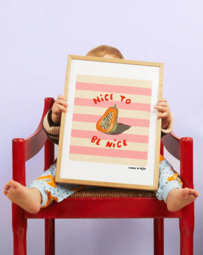 A baby sitting on a red chair, holding up a poster of a papaya in an oak frame in front of them.