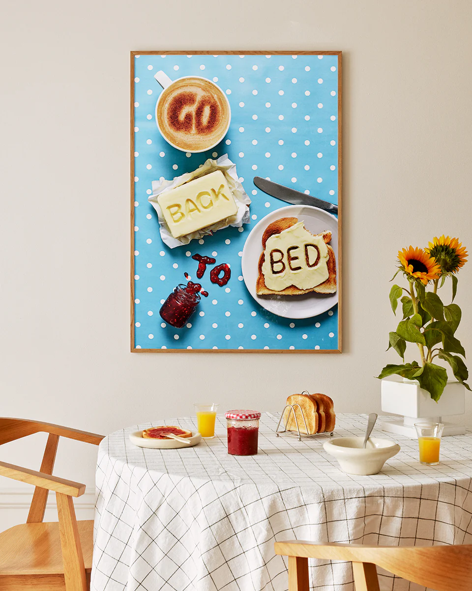 A cozy kitchen features a table with a checkered cloth and whimsical decor, including sunflowers in a vase. Above it hangs the Go Back to Bed Poster by Ellis Parrinder, adding playful charm with its toast and coffee theme.