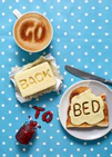 A mug of coffee spells 'GO' in froth, 'BACK' is intricately etched in butter, 'TO' is crafted from fruity jam, and 'bed' is toasted onto cheese toast. All arranged on a delightful blue tablecloth adorned with white polka dots.