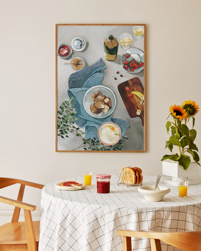 A Breakfast of Champions Poster featuring a breakfast table with bread and drinks hangs above a dining area. Below are toast, jam, juice, pancakes, a bowl, and a potted sunflower surrounded by wooden chairs.