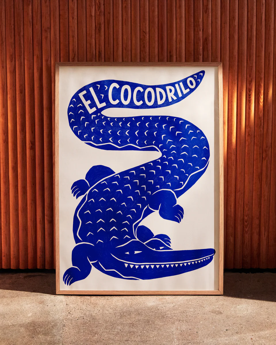 Framed El Cocodrilo Poster by Manne Jalilian features a stylized blue crocodile on a white background with El Cocodrilo elegantly inscribed above, set against a vertical wooden panel wall for added character.