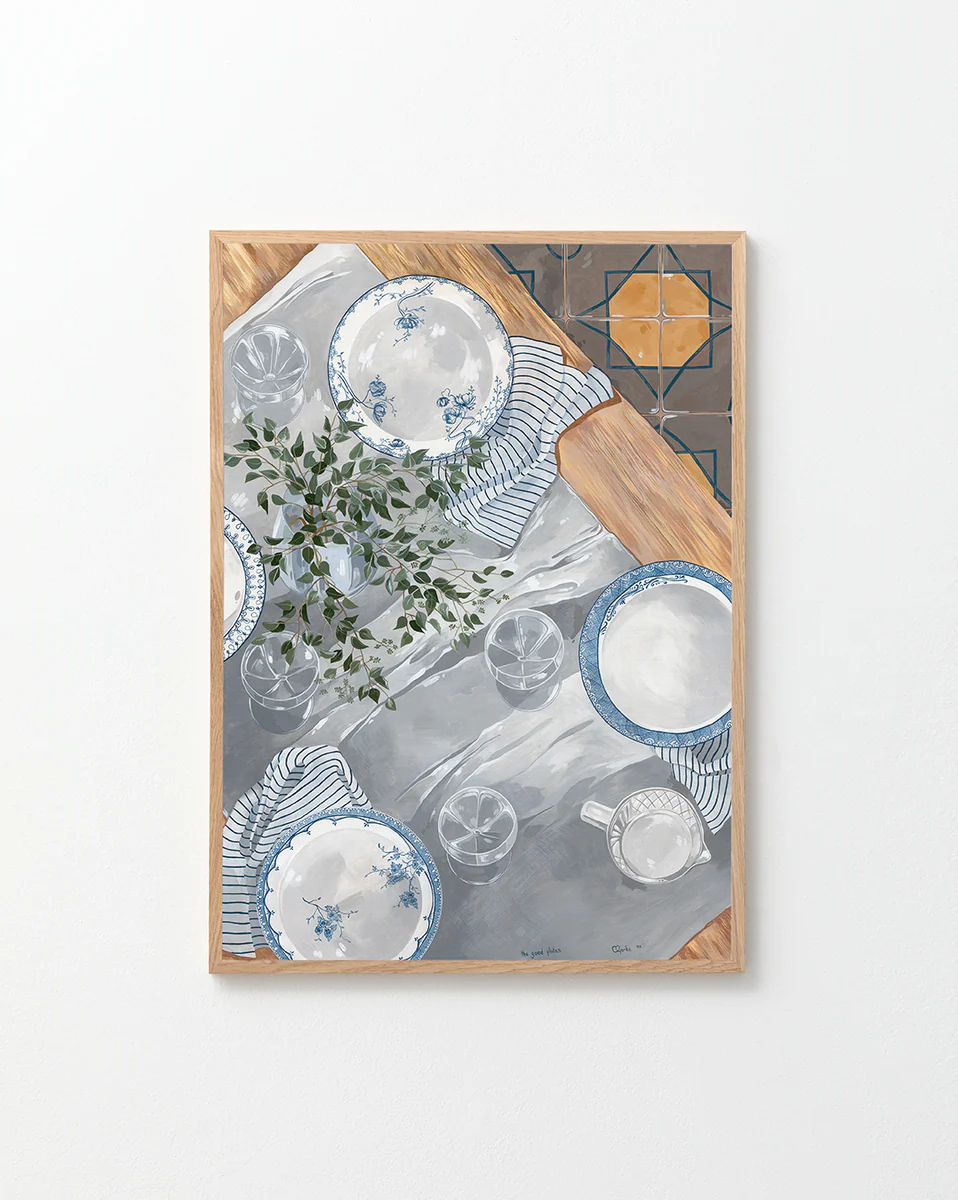 The Good Plates Poster displays a table setting with plates, striped napkins, glasses, and greenery on a white and gray surface. Wooden edges frame the scene, while a tiled corner pattern adds depth and charm.