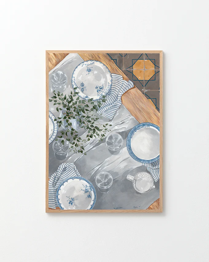 The Good Plates Poster displays a table setting with plates, striped napkins, glasses, and greenery on a white and gray surface. Wooden edges frame the scene, while a tiled corner pattern adds depth and charm.