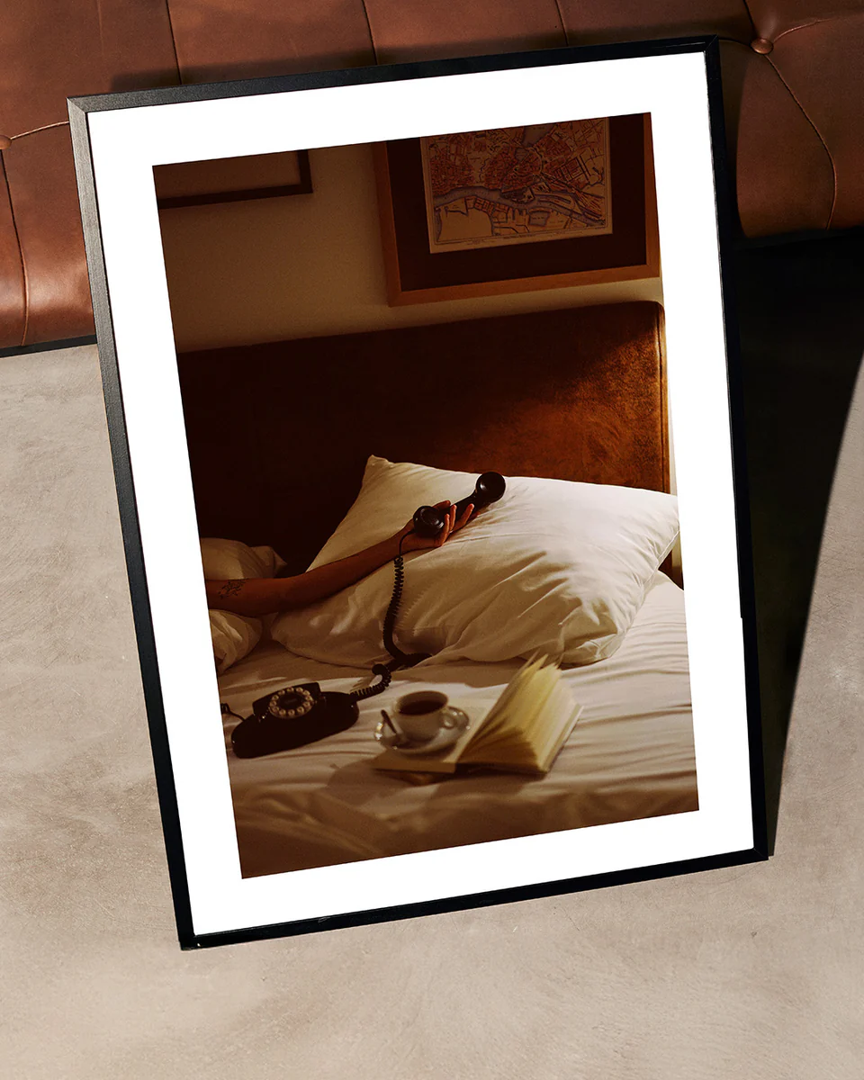 The Do Not Disturb Poster depicts a person reclining on a bed, cradling a vintage telephone. An open book and cup sit on the side table. With cozy, warm tones, it exudes the atmospheric essence of retro photography, lending an artistic touch to any space.