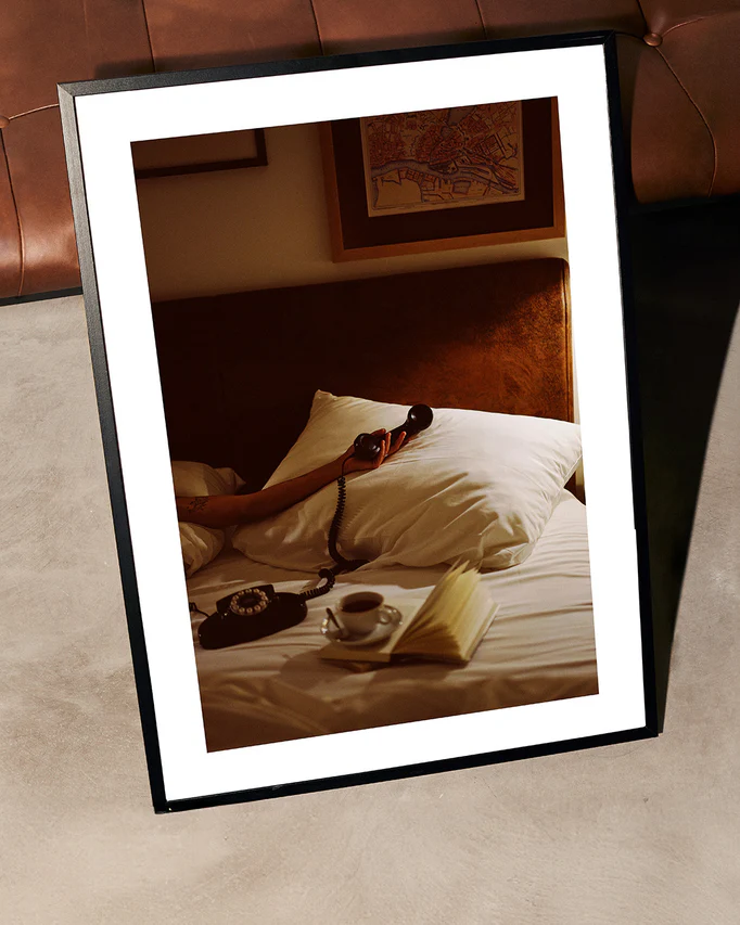 The Do Not Disturb Poster depicts a person reclining on a bed, cradling a vintage telephone. An open book and cup sit on the side table. With cozy, warm tones, it exudes the atmospheric essence of retro photography, lending an artistic touch to any space.