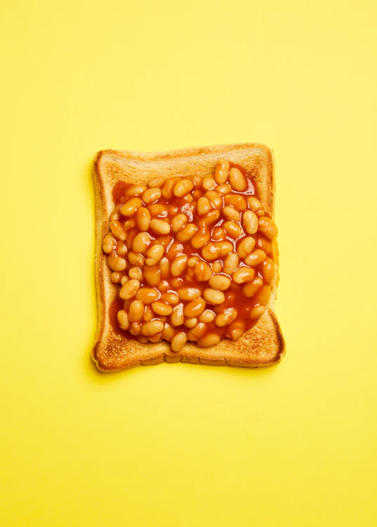 A still life photograph featuring baked beans on toast set against a vibrant yellow background.