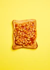A still life photograph featuring baked beans on toast set against a vibrant yellow background.