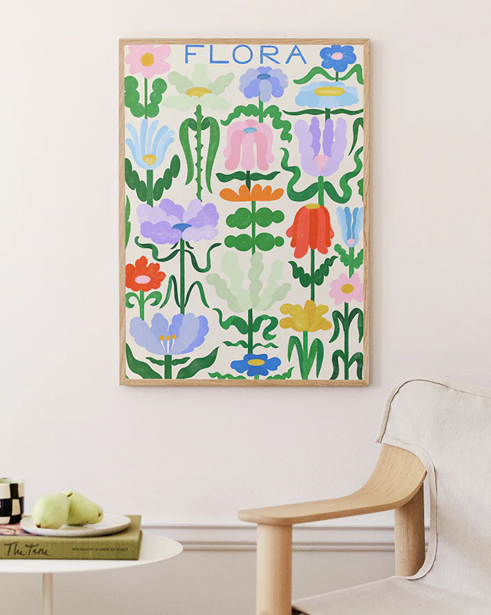 The Flora on White Poster showcases vibrant abstract flowers and leaves on a white wall, enhanced by a light wood chair and a round table with a book and green pears.