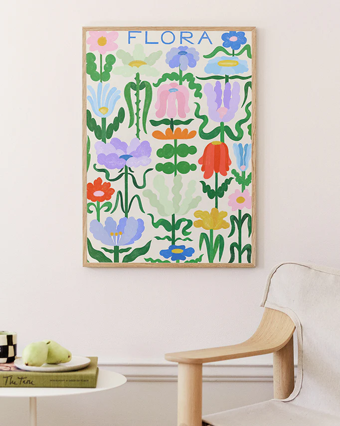 The Flora on White Poster showcases vibrant abstract flowers and leaves on a white wall, enhanced by a light wood chair and a round table with a book and green pears.