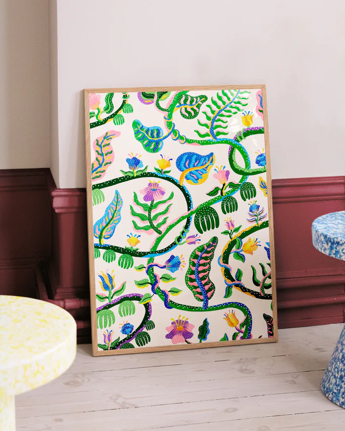 The Liana Poster by Manne Jalilian, featuring a vibrant design with green leaves, blue and orange flowers, and curving branches on a light backdrop, leans against the wall. Nearby, two round tables display textured surfaces in yellow with speckled patterns.