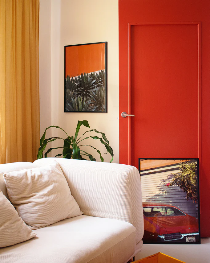 A cozy room includes a white couch with cream pillows, an orange-red door adorned with plant artwork, a floor plant beside a red car photo, and a vintage Pershing Square Red Poster above. A yellow curtain is on the left.