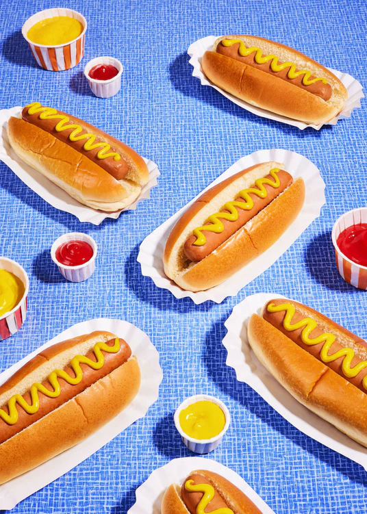 Still life photograph of perfectly grilled hot dogs adorned with mustard and served on a vibrant blue table cloth.