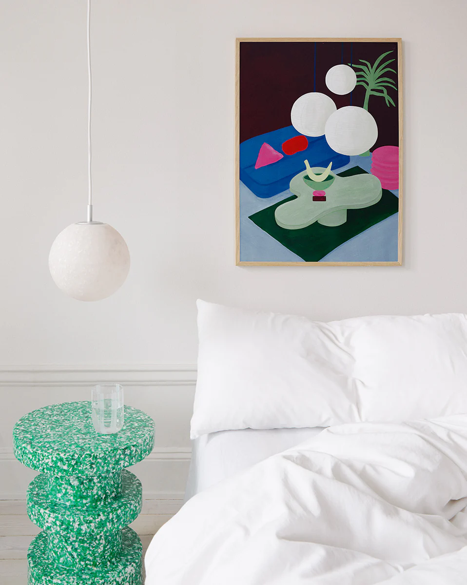 A minimalist bedroom features a white bed with fluffy pillows and a duvet, beside which stands a green terrazzo side table with a glass. The Colourful Living 01 Poster hangs above the bed, adding artistic flair, while a round pendant light graces the ceiling amid modern furnishings.