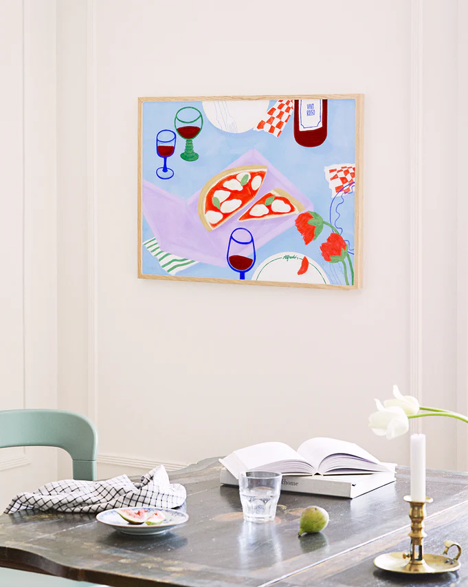 In this cozy kitchen scene, the Pizza for Three Poster by Frauke Schyroki displays a colorful pizza with wine glasses and a bottle. Below, a table is set with an open book, glass of water, candle, vase of white flowers, checkered cloth, and small plate to complete the charming tableau.
