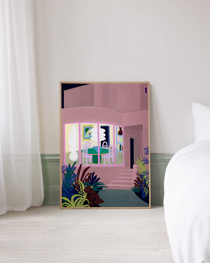 The Lighted Windows 01 Poster, featuring a modern pink building with large windows and surrounding plants, leans against a wall in a room with light-colored walls, a white bed, and sheer curtains.