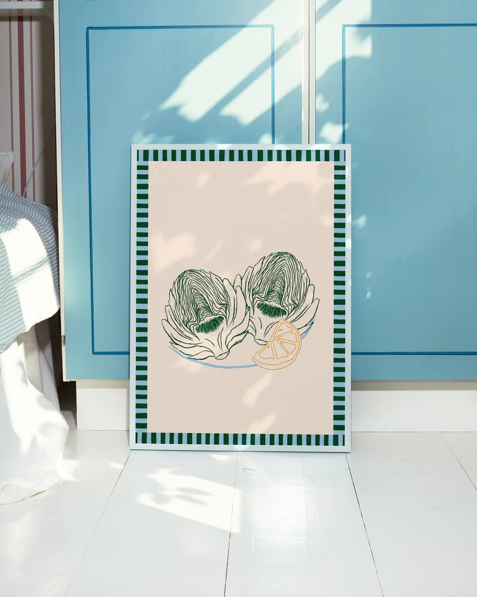 The Les Artichauts Poster features two artichokes and lemon slices with a beige background and green striped border. It leans against blue cabinet doors on a sunlit white wooden floor, bringing the charm of a French market to your home decor.