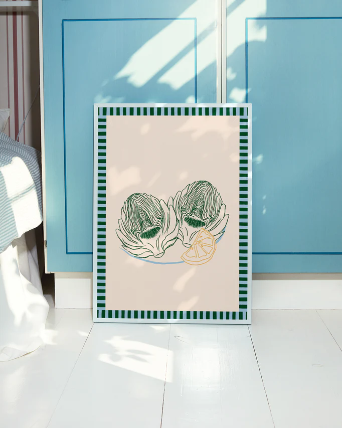 The Les Artichauts Poster features two artichokes and lemon slices with a beige background and green striped border. It leans against blue cabinet doors on a sunlit white wooden floor, bringing the charm of a French market to your home decor.