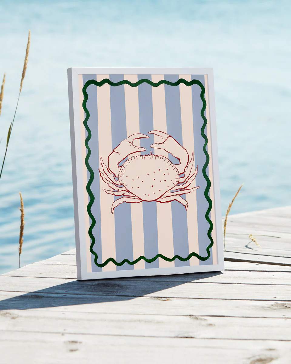 Le Crabe Poster embodies a coastal vibe with its illustration of a red crab on vertical blue and gray stripes, bordered by a green wave. Displayed against a wooden deck by water, it features tall grass gently swaying in the background.