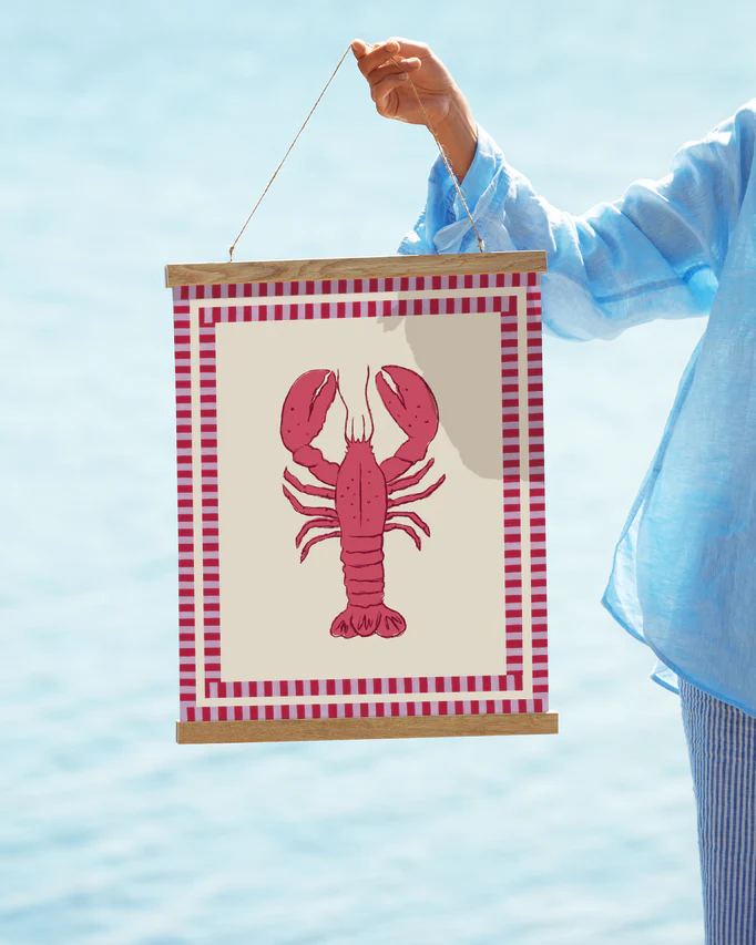 A person in a light blue shirt holds the Le Stripy Homard Poster, a collage art piece with a red lobster illustration, framed with a checkered red and white border. The backdrop is a blurred blue, evoking the essence of water.