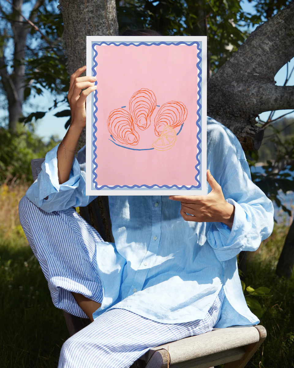 A person outdoors, wearing a light blue shirt and striped pants, holds the vibrant *Plateau dhuîtres Poster*, which is pink with illustrations of three oysters and a lemon slice, bordered by a wavy blue line. Trees and greenery surround them as the poster obscures their face.
