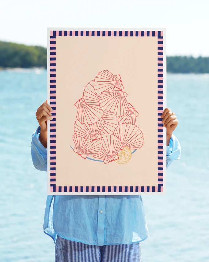 In a blue shirt and striped pants, someone holds the Saint-Jacques du Marché Poster, featuring abstract red line art of seashells in collage style with a small yellow lemon. A body of water and trees appear in the background, while the poster obscures their face.
