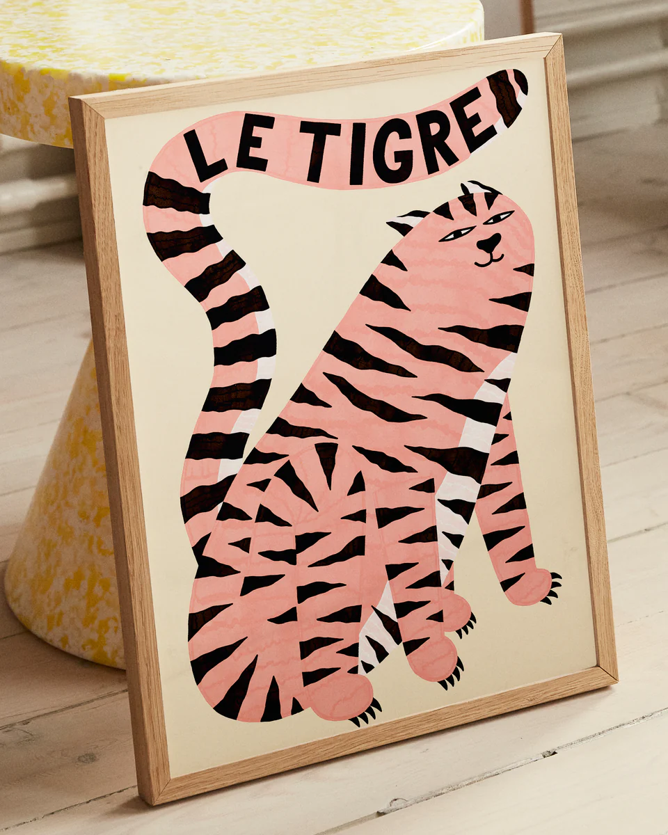 The Le Tigre White Poster showcases a stylized pink tiger illustration with black stripes, featuring the title LE TIGRE boldly on its tail. Leaning against a table with a yellow and white pattern, it adds wild sophistication to the light wooden flooring.