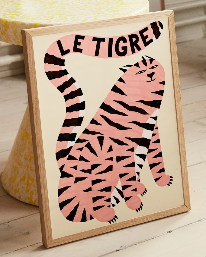 The Le Tigre White Poster showcases a stylized pink tiger illustration with black stripes, featuring the title LE TIGRE boldly on its tail. Leaning against a table with a yellow and white pattern, it adds wild sophistication to the light wooden flooring.
