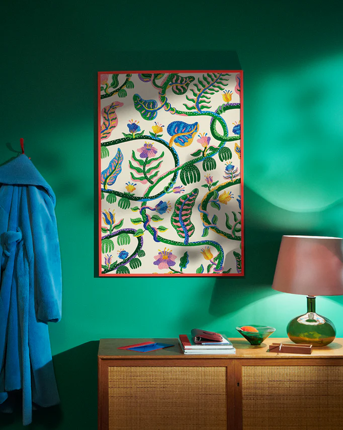 A vibrant room with green walls features the colorful Liana Poster by Manne Jalilian in a red frame. Below, a wooden console holds books and decor. A blue coat hangs on a hook, while a lamp with a pink shade casts warm light.