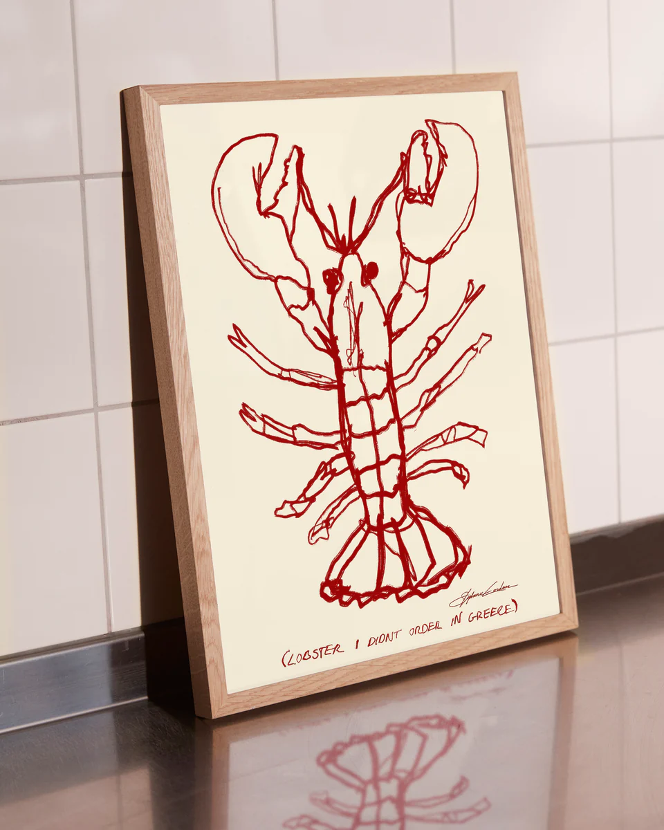 A Lobster Poster, featuring a red lobster drawing on beige with the caption (LOBSTER, I DONT ORDER IN GREECE), adorns the kitchen countertop against elegantly tiled white walls.