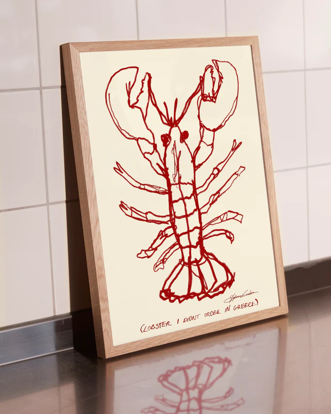 A Lobster Poster, featuring a red lobster drawing on beige with the caption (LOBSTER, I DONT ORDER IN GREECE), adorns the kitchen countertop against elegantly tiled white walls.