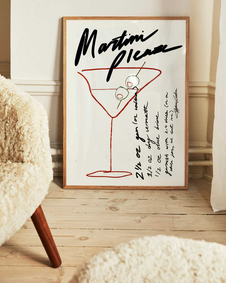 The Martini Please Poster, displayed on a wooden floor, features a stylish illustration of a martini glass with olives alongside handwritten measurements and instructions for a classic martini recipe. A fuzzy chair in the foreground enhances the cozy ambiance.