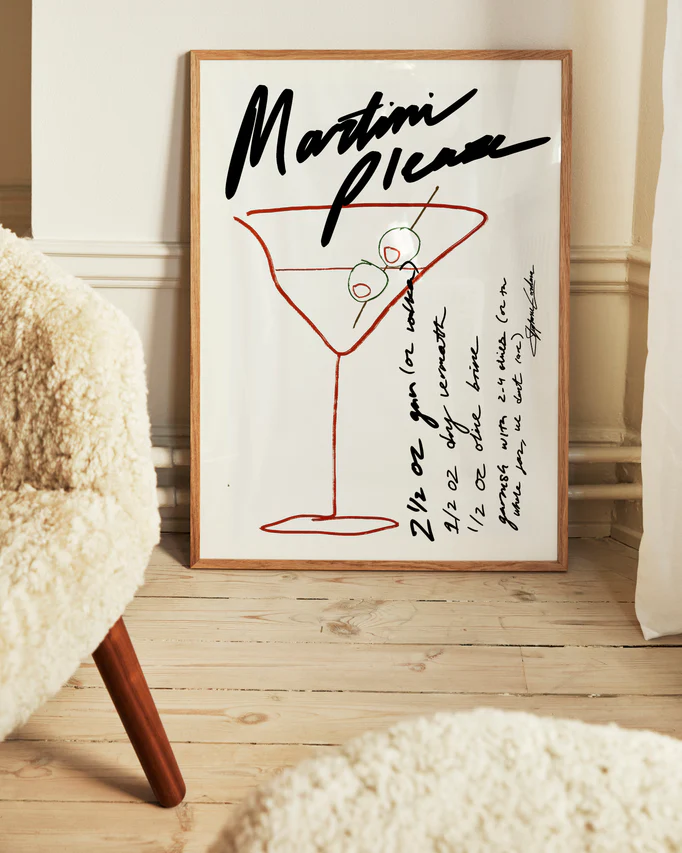 The Martini Please Poster, displayed on a wooden floor, features a stylish illustration of a martini glass with olives alongside handwritten measurements and instructions for a classic martini recipe. A fuzzy chair in the foreground enhances the cozy ambiance.