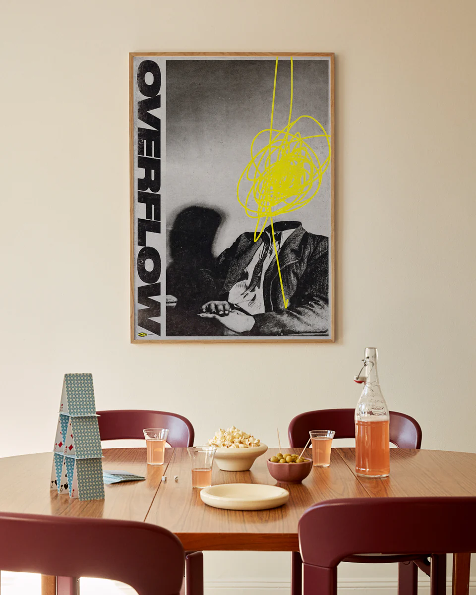 A dining table with maroon chairs features stacked cards, glasses of drinks, a bottle, popcorn, and olives. Above it hangs the Overflow Poster by Meinhard Taxer with the word OVERFLOW and a yellow scribble on its black-and-white design.