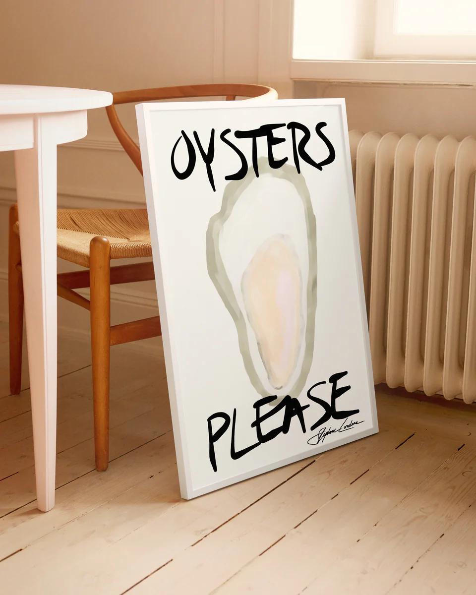 The Oysters Please Poster leans against a wall, featuring a watercolor oyster with big black letters. Its on a wooden floor next to a chair by a white table, bathed in light from the nearby window.