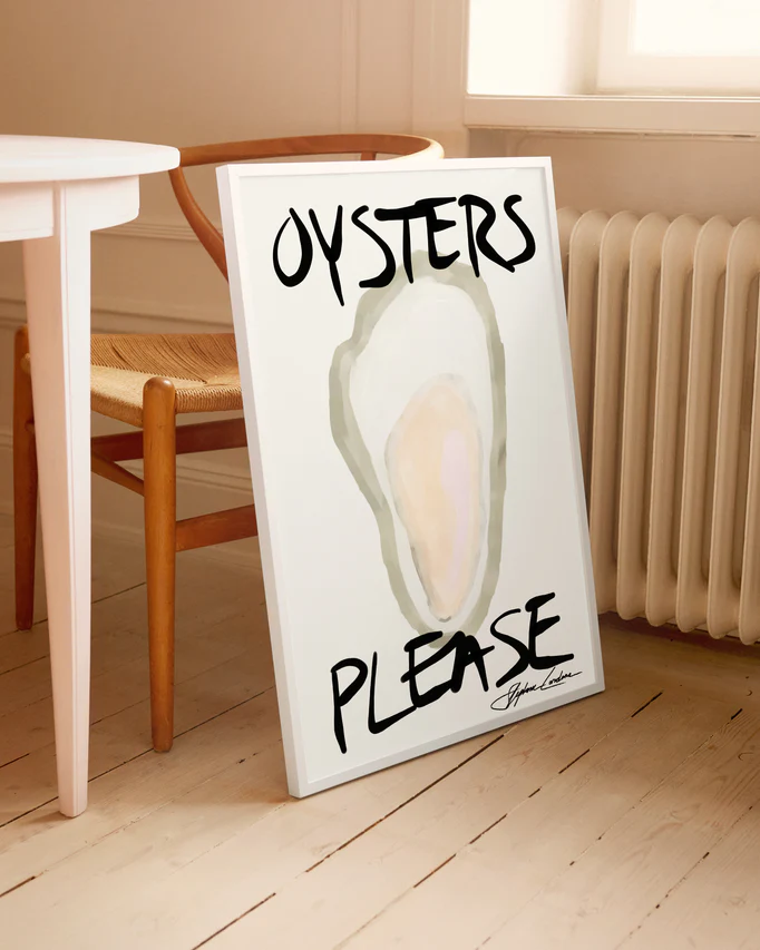 The Oysters Please Poster leans against a wall, featuring a watercolor oyster with big black letters. Its on a wooden floor next to a chair by a white table, bathed in light from the nearby window.