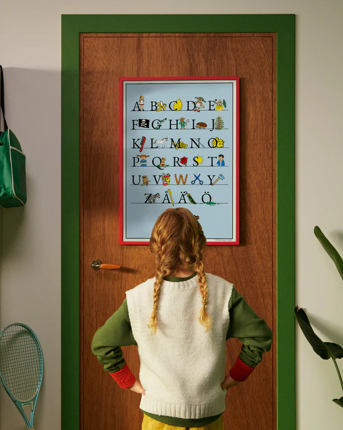 A child standing in front of a door looking at a Pippi Longstocking Swedish alphabet poster A–Ö.