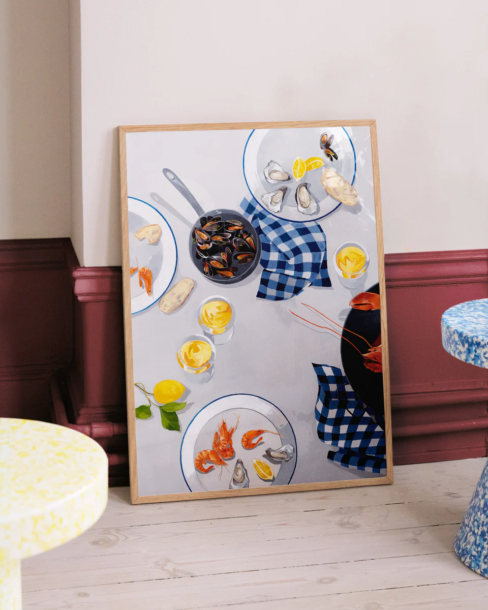 The Break Out of the Shell I Poster by Cat Gerke elegantly rests on the floor, showcasing a seafood-themed table setting with mussels in a pot and shellfish, complete with lemon wedges and a blue checkered napkin, leaning against a two-toned wall above a wooden floor.
