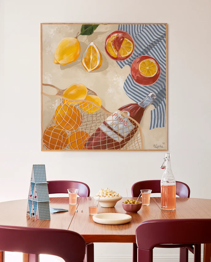 A dining area with a wooden table set for snacks, featuring popcorn, olives, drinks in glasses, and a bottle. Behind the table is the Pimms Afternoon Poster by Cat Gerke, showcasing lemons, oranges, a net with a bottle, and a blue-striped cloth.