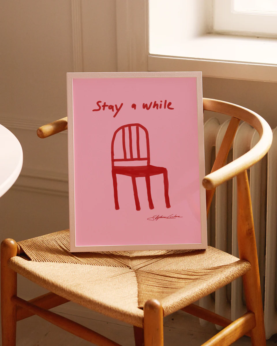 The Stay a While Poster, with its red chair illustration and text, rests on a woven wooden chair, adding charm to the decor. Natural light from a nearby window softly illuminates the scene.