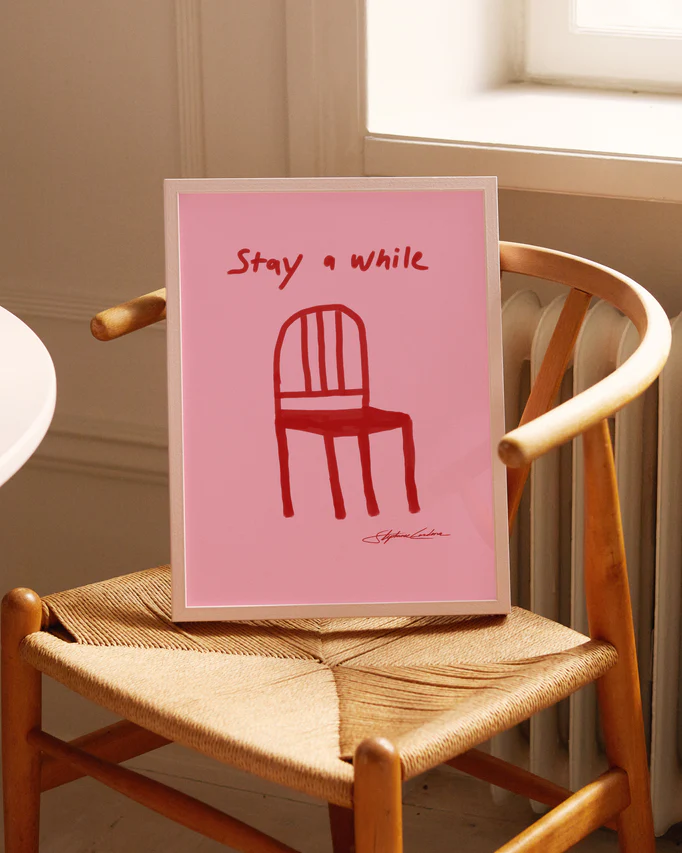 The Stay a While Poster, with its red chair illustration and text, rests on a woven wooden chair, adding charm to the decor. Natural light from a nearby window softly illuminates the scene.