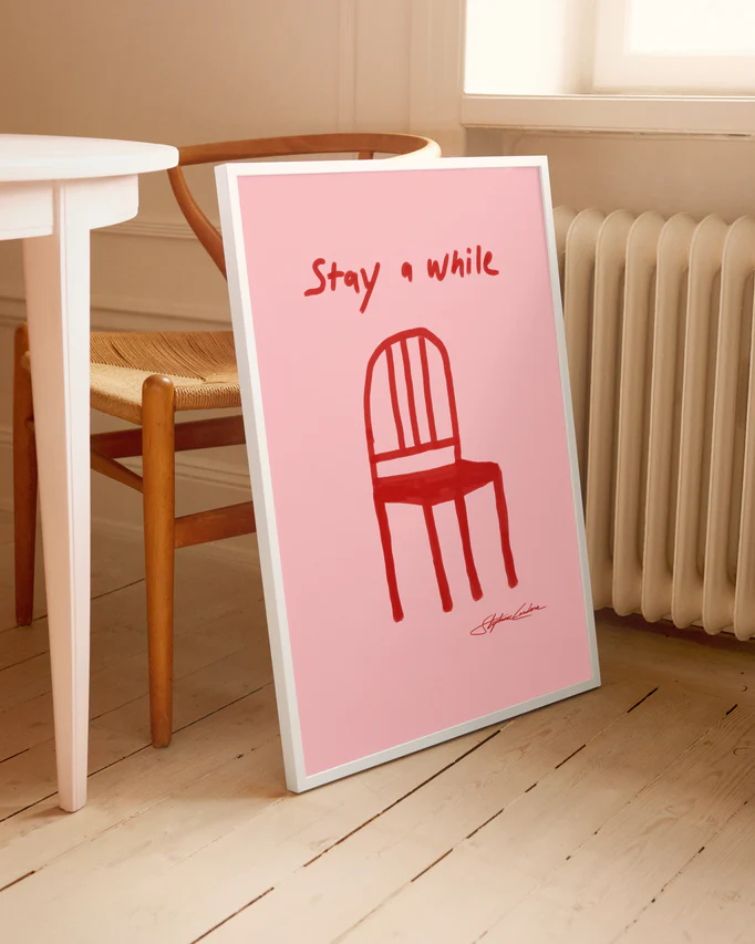 The Stay a While Poster, showcasing a red chair illustration and text, leans against the wall on a light wooden floor. Its beside a wooden chair and white table, creating an inviting home decor scene as soft light from a nearby window enhances its serenity.