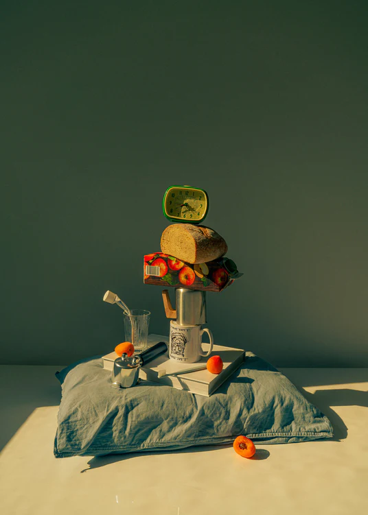 A still life featuring various breakfast elements including a slice of bread, apple juice, coffee mug, teapot, with a pillow underneath and an alarm clock on top.