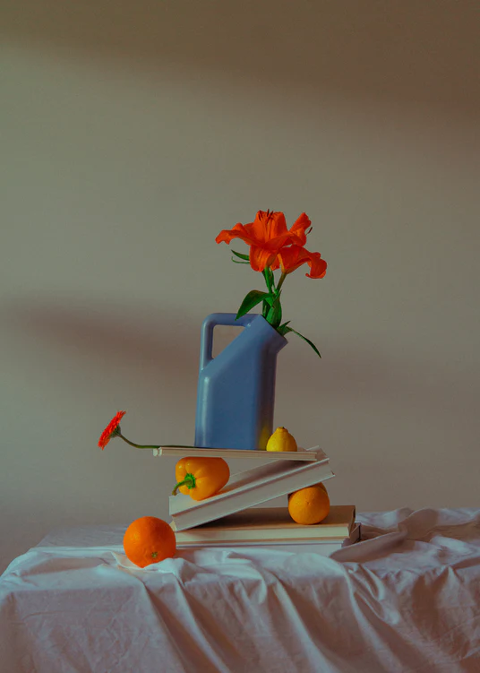 The Hanging in There Poster showcases a still life with a blue pitcher holding vibrant orange flowers, exuding warmth amid winters stark simplicity. Its set on books, surrounded by scattered oranges and a lemon, all atop a white cloth, capturing Berlins artistic essence.