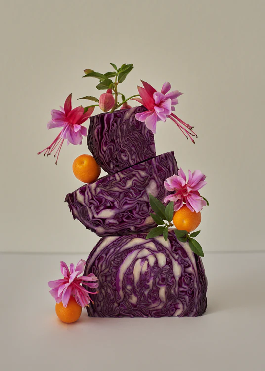 A still life composition of red cabbages balanced on top of each other, adorned with pink flowers, set against a white background.