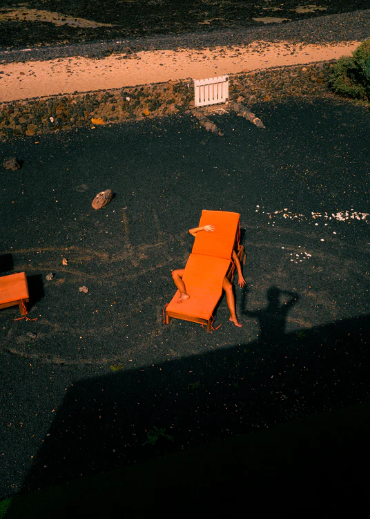 A person playfully poses under an orange table on dark gravel, one arm and leg extended, while the photographers shadow blends with scattered vegetation and rocks. Its a scene reminiscent of The Short Romantic Story Between a Chair and a Shadow Poster.