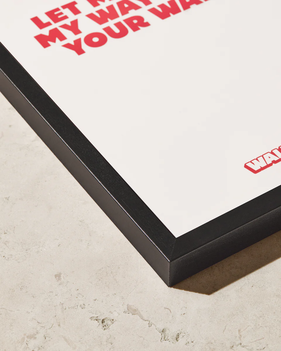 Close-up of a framed artwork on a beige textured surface. The Frame Black provides a classic look, showcasing bold red text: LET MY WAR... YOUR WAR... with WAR... in the bottom right corner, beautifully preserved behind high-quality acrylic glass.