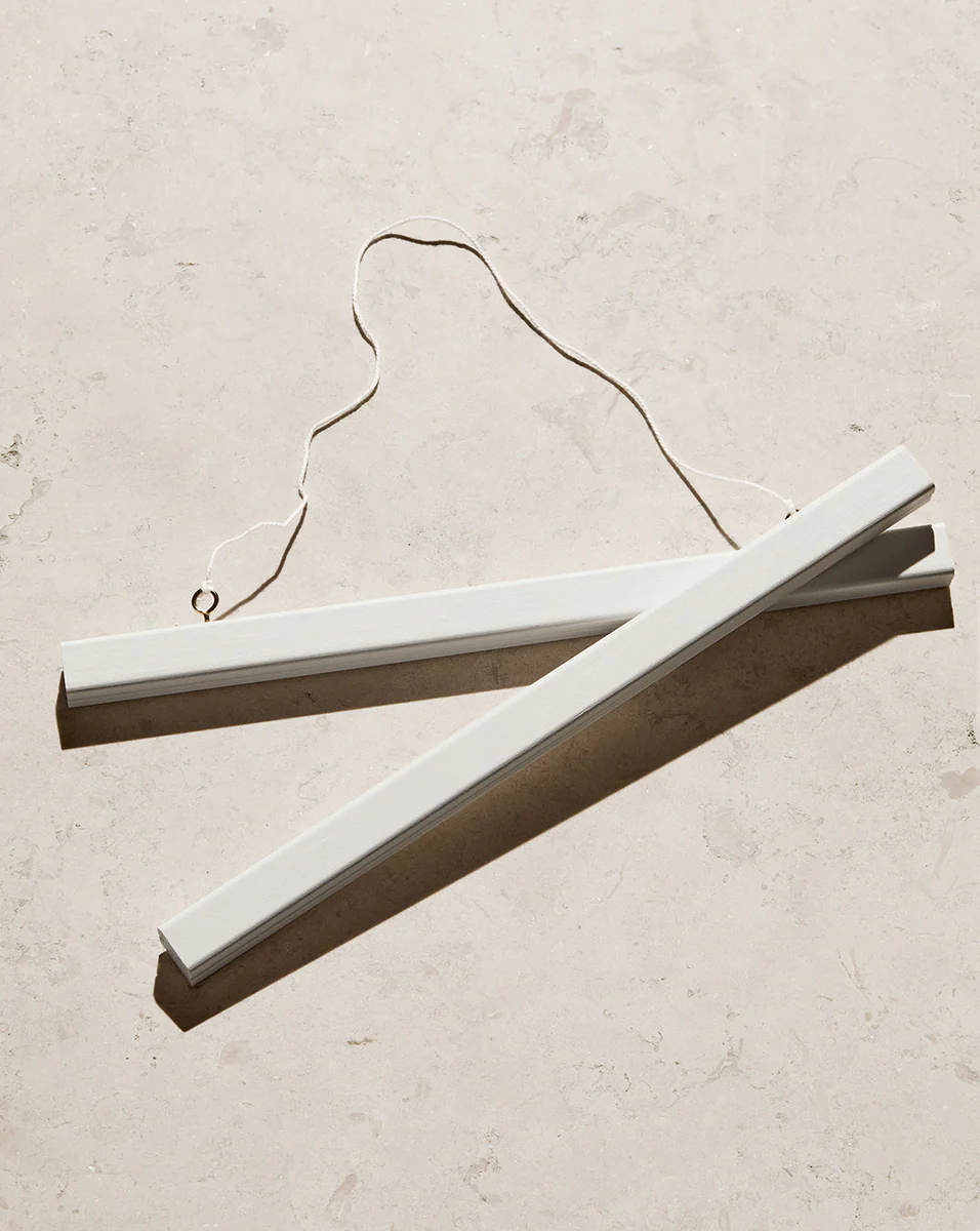 Two Poster hanger White pieces crafted from solid pine, complete with an attached string, rest on a light, textured surface. Sunlight casts soft shadows that highlight their minimalist design and the discreet magnets that effortlessly secure your art.