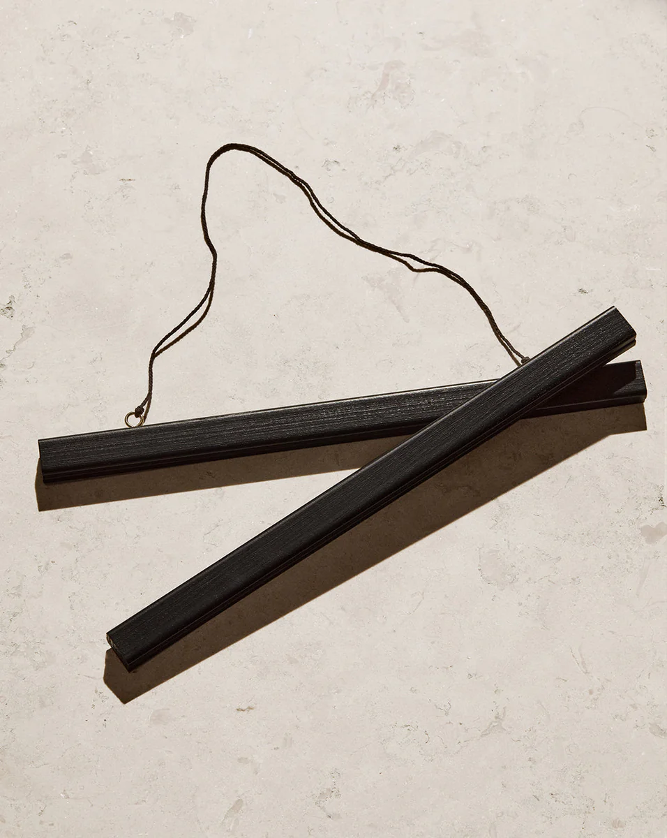 A Poster hanger Black with two sleek black wooden sticks and a string attached lays diagonally on a light marble surface, showcasing its thin rectangular shape and looped string design.